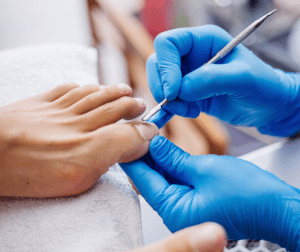 Close-up of a nail technician in blue gloves using a cuticle tool on a client's nail during a manicure/pedicure.
